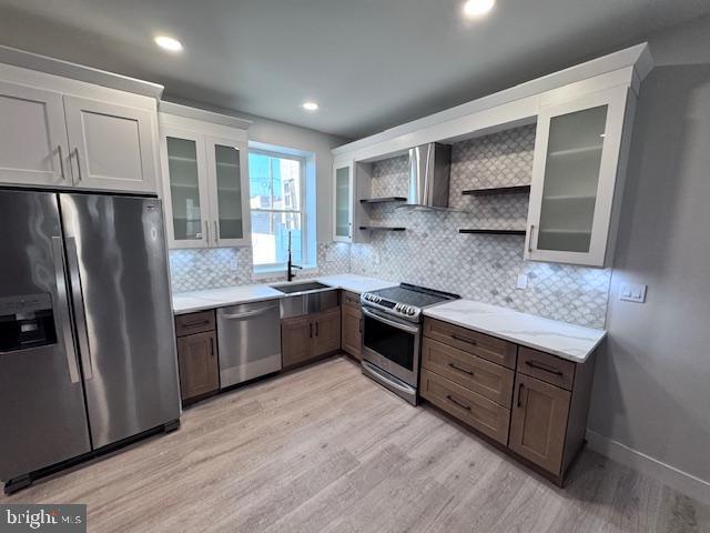 kitchen with stainless steel appliances, open shelves, a sink, light wood-style floors, and wall chimney exhaust hood