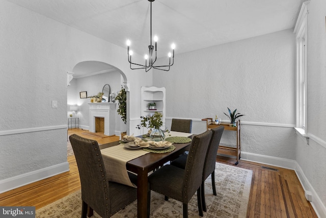 dining room featuring a notable chandelier, a fireplace with raised hearth, wood finished floors, arched walkways, and a textured wall