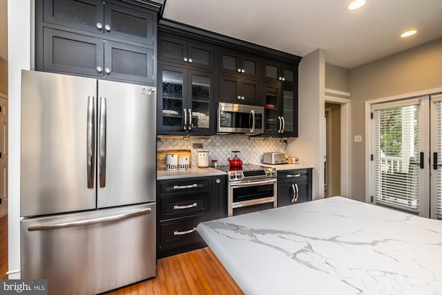 kitchen with dark cabinetry, stainless steel appliances, glass insert cabinets, tasteful backsplash, and light wood-type flooring