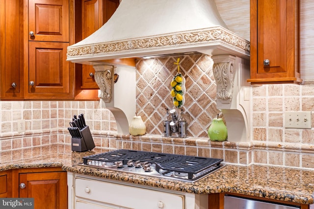 kitchen with custom range hood, backsplash, light stone countertops, brown cabinetry, and stainless steel gas stovetop