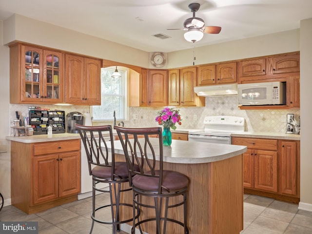 kitchen featuring white appliances, visible vents, glass insert cabinets, under cabinet range hood, and a kitchen bar