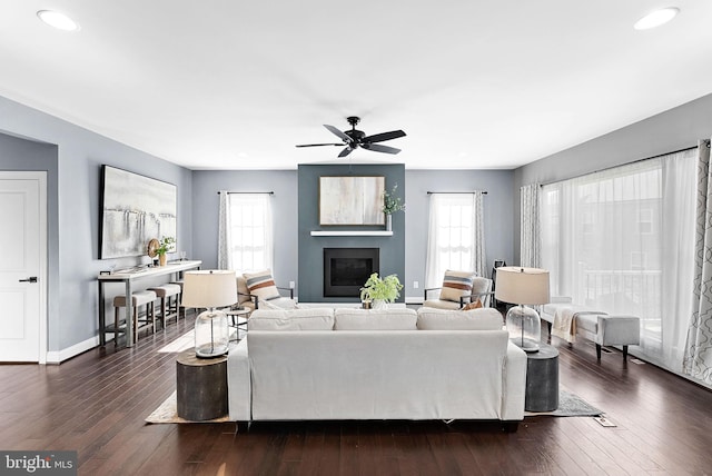 living room featuring ceiling fan, a fireplace, baseboards, and dark wood-style flooring