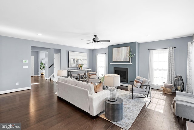 living area featuring dark wood-style floors, baseboards, a ceiling fan, a fireplace, and stairs