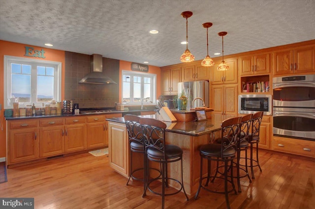 kitchen with light wood finished floors, wall chimney exhaust hood, appliances with stainless steel finishes, and a kitchen breakfast bar