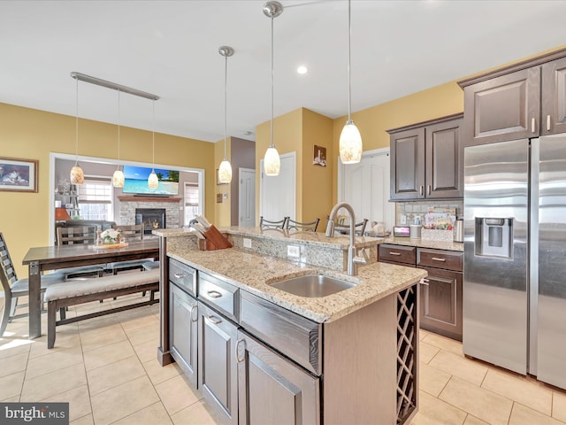 kitchen featuring light tile patterned floors, hanging light fixtures, a stone fireplace, stainless steel refrigerator with ice dispenser, and a sink