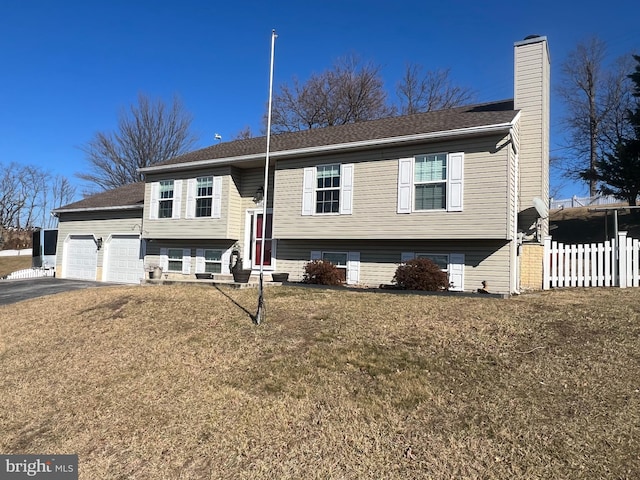 bi-level home featuring aphalt driveway, an attached garage, fence, a chimney, and a front yard