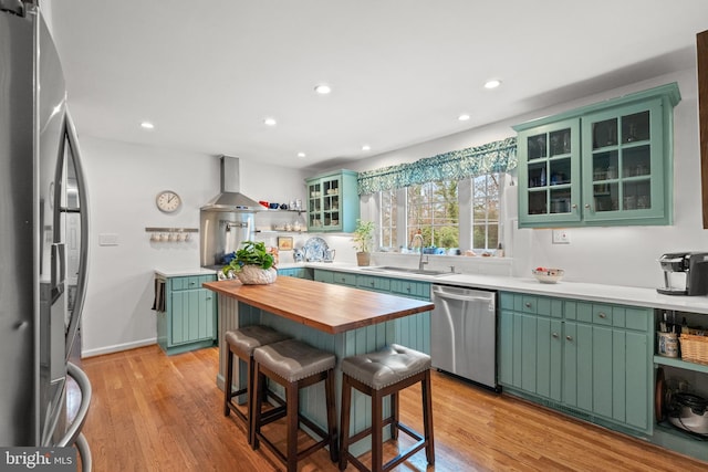 kitchen with wooden counters, green cabinets, appliances with stainless steel finishes, a sink, and wall chimney exhaust hood