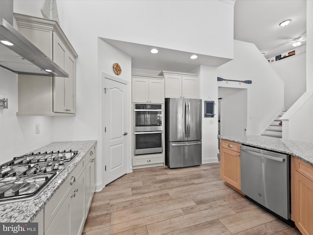 kitchen featuring light stone counters, light wood-style floors, appliances with stainless steel finishes, and wall chimney range hood