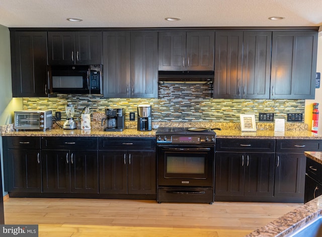 kitchen featuring under cabinet range hood, light wood-type flooring, light stone countertops, and black appliances