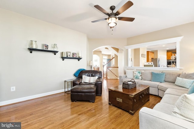 living room with ceiling fan, baseboards, stairway, light wood-style floors, and arched walkways