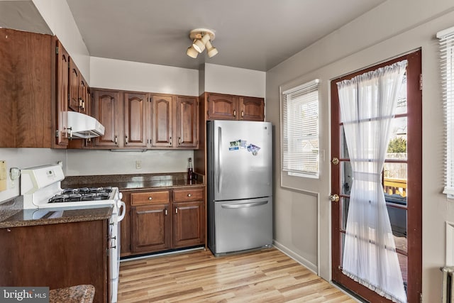 kitchen with under cabinet range hood, white range with gas stovetop, freestanding refrigerator, light wood finished floors, and dark countertops