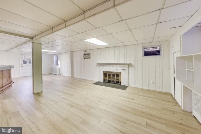 finished basement with light wood-type flooring, an AC wall unit, a drop ceiling, and a fireplace