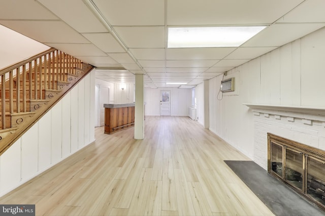 finished below grade area featuring stairway, a drop ceiling, a brick fireplace, light wood-type flooring, and a wall mounted air conditioner