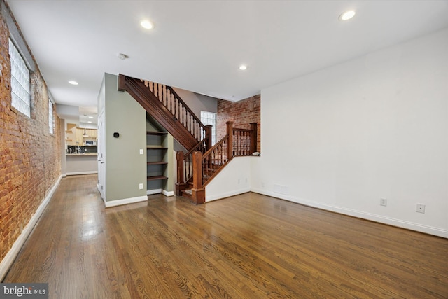 unfurnished living room featuring stairway, baseboards, wood finished floors, and brick wall