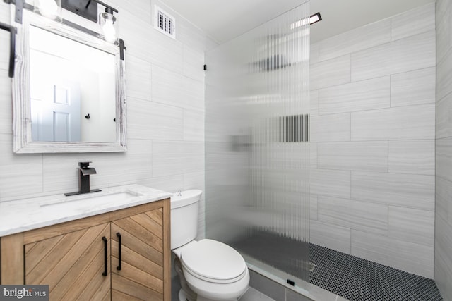 full bathroom featuring visible vents, toilet, a tile shower, vanity, and tile walls