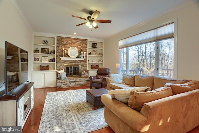 living room featuring built in features, crown molding, a fireplace, a ceiling fan, and wood finished floors