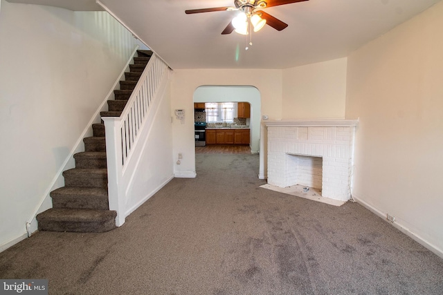 unfurnished living room with arched walkways, carpet flooring, a ceiling fan, stairs, and a brick fireplace