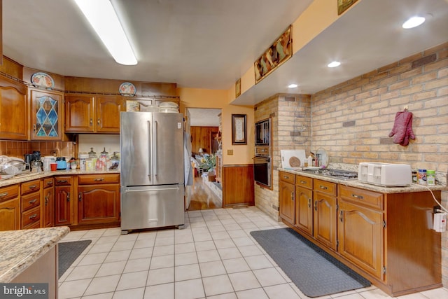 kitchen with a wainscoted wall, light stone counters, appliances with stainless steel finishes, brick wall, and brown cabinetry