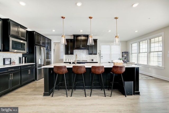 kitchen featuring a kitchen island with sink, stainless steel appliances, light countertops, light wood-style floors, and backsplash