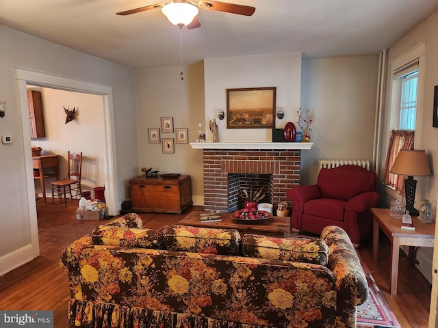 living area featuring baseboards, radiator, ceiling fan, wood finished floors, and a brick fireplace