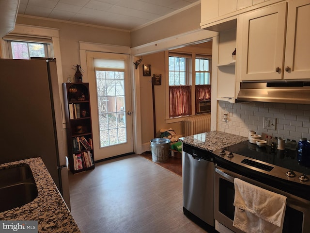 kitchen featuring radiator heating unit, appliances with stainless steel finishes, dark stone countertops, crown molding, and under cabinet range hood