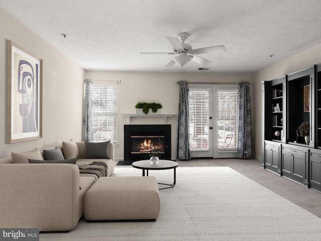 carpeted living area featuring visible vents, a ceiling fan, a textured ceiling, a warm lit fireplace, and french doors