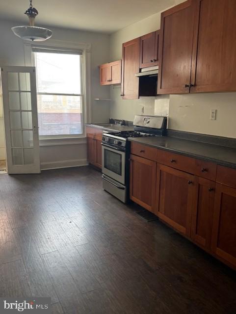 kitchen featuring under cabinet range hood, dark wood-style flooring, stainless steel gas stove, brown cabinetry, and dark countertops