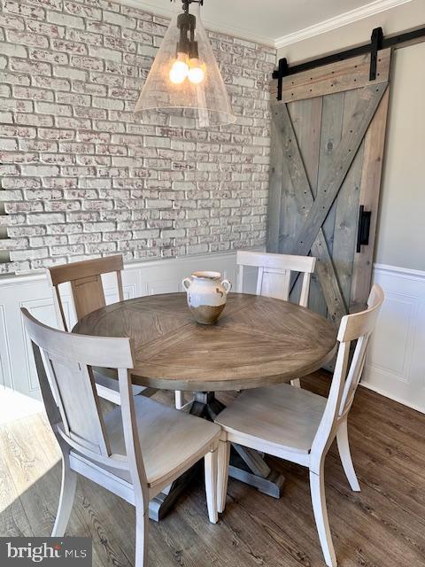 dining room with a barn door, a wainscoted wall, brick wall, wood finished floors, and ornamental molding