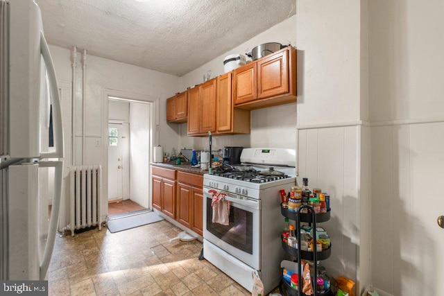 kitchen with a textured ceiling, a wainscoted wall, white appliances, radiator, and stone finish flooring