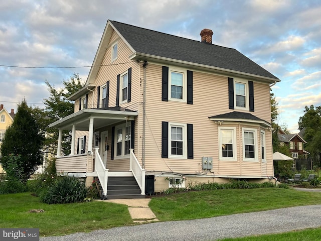 view of front of home with a front yard, covered porch, roof with shingles, and a chimney