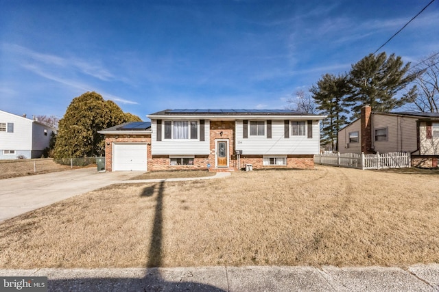 raised ranch featuring solar panels, concrete driveway, fence, a front lawn, and brick siding