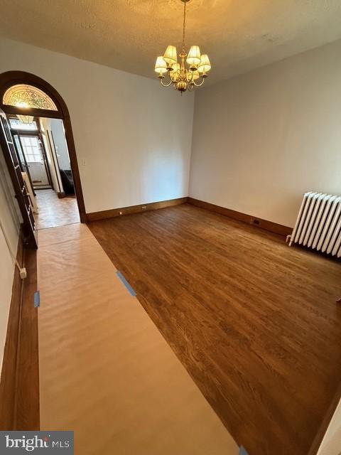 empty room featuring baseboards, arched walkways, radiator heating unit, wood finished floors, and a chandelier