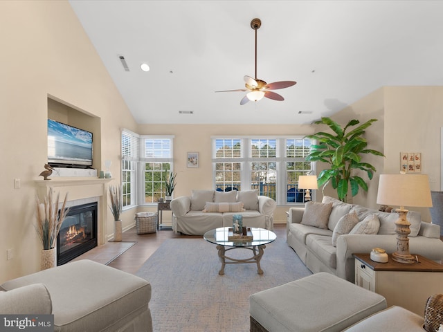 living room featuring a ceiling fan, wood finished floors, visible vents, high vaulted ceiling, and a glass covered fireplace