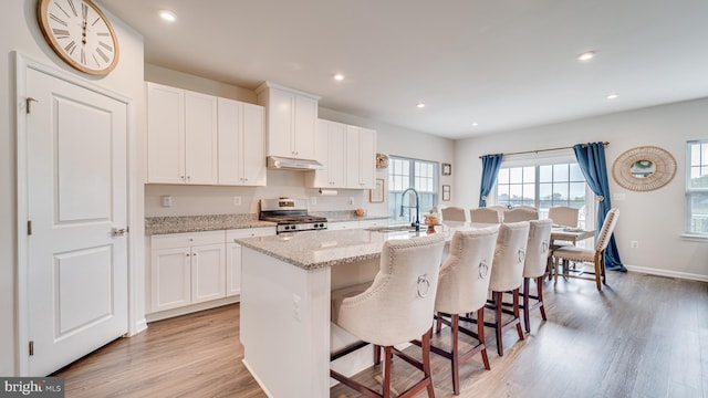 kitchen with light stone counters, a kitchen island with sink, under cabinet range hood, white cabinets, and stainless steel range with gas stovetop