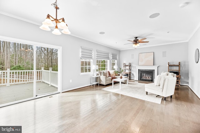 living area featuring baseboards, a glass covered fireplace, wood finished floors, crown molding, and ceiling fan with notable chandelier