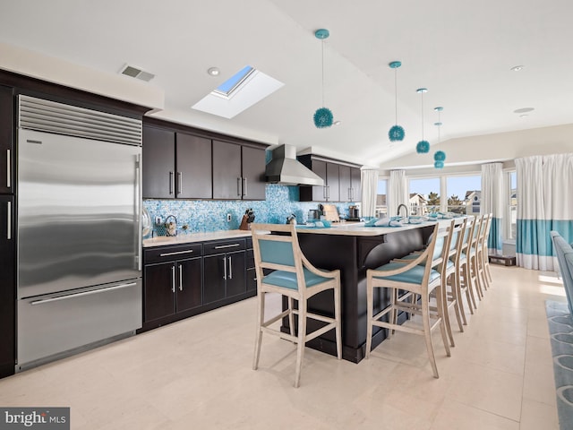 kitchen featuring vaulted ceiling with skylight, visible vents, light countertops, wall chimney exhaust hood, and stainless steel built in refrigerator