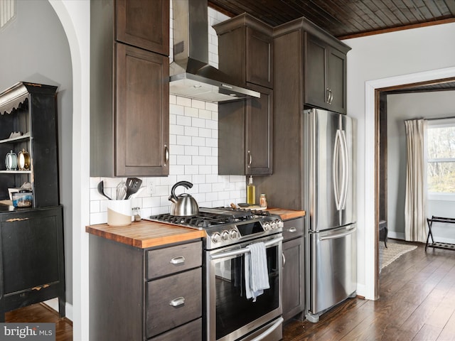 kitchen featuring wooden counters, backsplash, appliances with stainless steel finishes, dark brown cabinets, and wall chimney exhaust hood