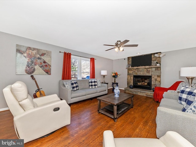 living area with ceiling fan, baseboards, a stone fireplace, and wood finished floors