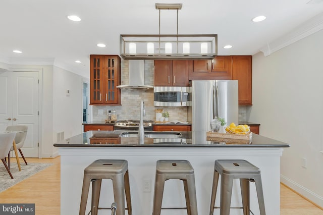 kitchen with stainless steel appliances, ornamental molding, brown cabinets, wall chimney exhaust hood, and dark countertops