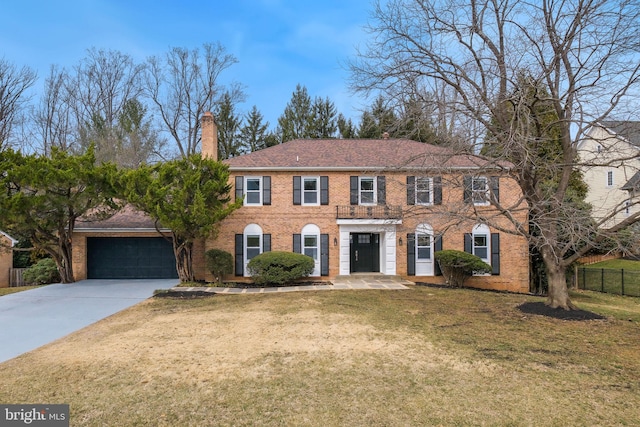 colonial house featuring brick siding, a chimney, concrete driveway, fence, and a front lawn