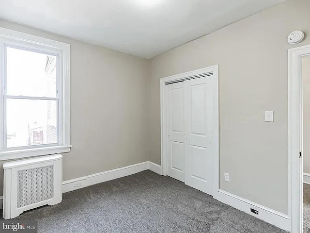 unfurnished bedroom with a closet, baseboards, dark carpet, and radiator heating unit