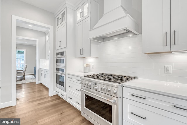 kitchen with light wood finished floors, appliances with stainless steel finishes, light countertops, premium range hood, and white cabinetry