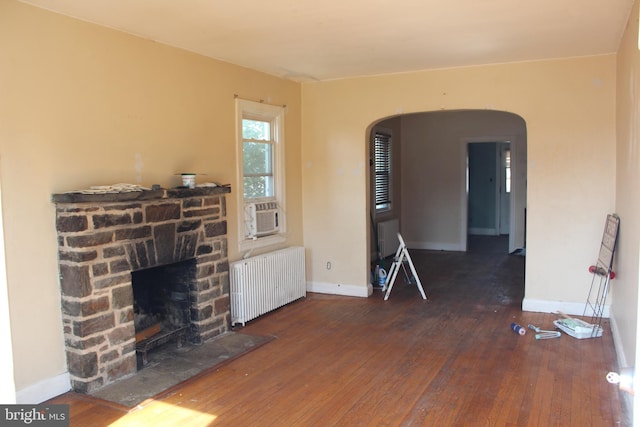 unfurnished living room with baseboards, arched walkways, radiator, hardwood / wood-style floors, and a stone fireplace