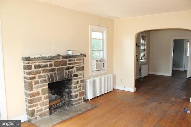 unfurnished living room featuring radiator heating unit, arched walkways, hardwood / wood-style floors, and a stone fireplace