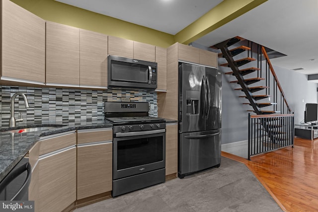 kitchen featuring light wood finished floors, backsplash, appliances with stainless steel finishes, a sink, and dark stone counters