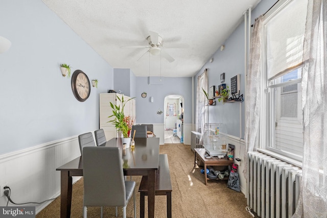 carpeted dining room featuring arched walkways, a textured ceiling, a wainscoted wall, a ceiling fan, and radiator heating unit