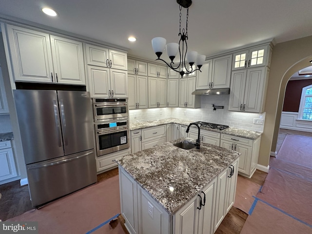 kitchen with arched walkways, under cabinet range hood, stainless steel appliances, a sink, and tasteful backsplash