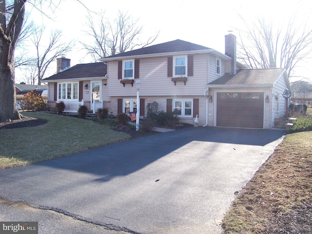 split level home featuring aphalt driveway, brick siding, a chimney, a garage, and a front lawn