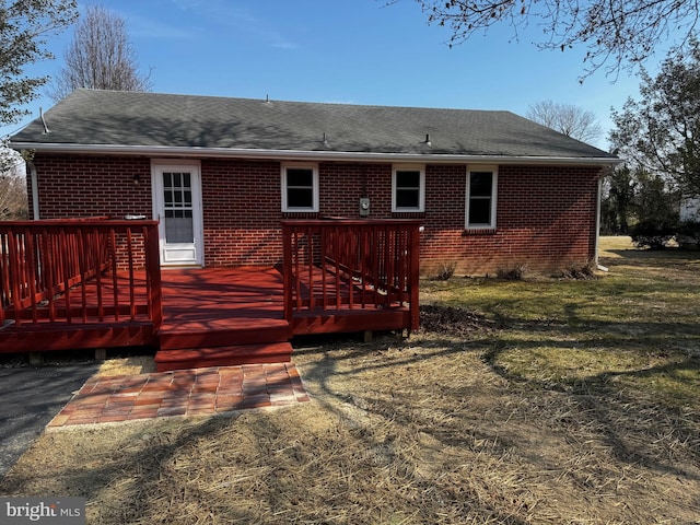 rear view of property featuring a deck, roof with shingles, brick siding, and a lawn