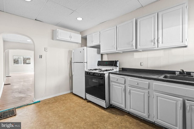 kitchen with arched walkways, a drop ceiling, white appliances, a sink, and dark countertops